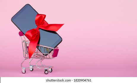 Shopping Cart With Black Mobile Phone With Bright Red Ribbon On Pink Background. Copy Space.