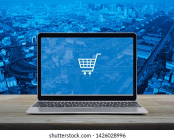 Shopping cart flat icon with modern laptop computer on wooden table over city tower, street, expressway and skyscraper, Business shop online concept