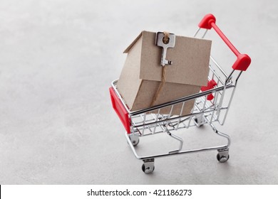 Shopping Cart With Model Of Cardboard House On Gray Background, Buying A New Home Or Sale Of Real Estate Concept