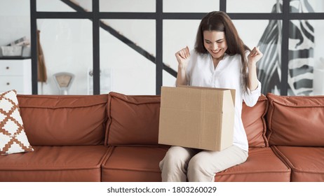 Shopping Delivery Concept. Happy And Excited Young Woman Sitting On Couch Wearing In Casual Clothes, Unpacking Carton Box, Raised Hand Up And Smiling Wide