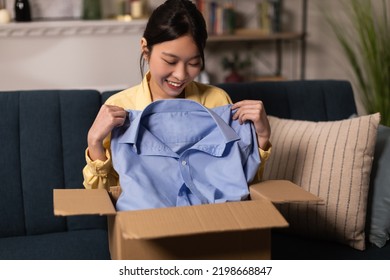 Shopping And Delivery. Joyful Chinese Lady Unpacking Cardboard Box Holding Blue Shirt Receiving Parcel From Shop Sitting On Couch At Home. Ecommerce Service, Contented Customer Concept