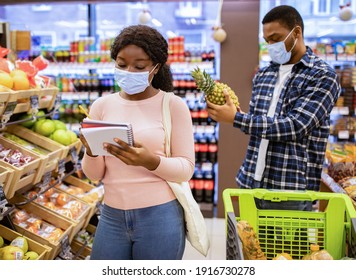 Shopping During Covid-19 Outbreak. Young Black Lady In Face Mask And Her Boyfriend Buying Fruits, Checking Grocery List At Mall. Millennial Couple Purchasing Food Products At Supermarket