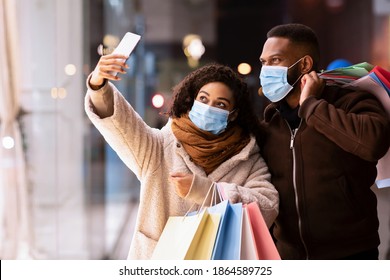 Shopping During Quarantine And Pandemic. Happy african american couple wearing medical face masks and taking selfie near mall in the evening, holding shopper bags, smiling at camera of cellphone