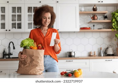 Shopping Economy. Happy Black Woman In Kitchen Checking Bill After Grocery Shopping, Smiling African American Female Standing Near Table, Counting Spends, Enjoying Affordable Prices For Food - Powered by Shutterstock