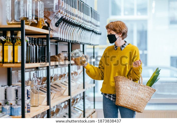 Shopping, food, sale, consumerism concept. Blond\
short haired woman in face protective mask holds glass jar with\
soya, buying at grocery store Reusable wicker basket for shopping.\
Zero waste concept.