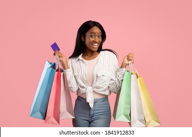 Shopping Frenzy Concept. Smiling Black Lady With Gift Bags And Credit Card Buying Holiday Presents On Pink Background. Excited Shopaholic With Bunch Of Gifts And Bank Card