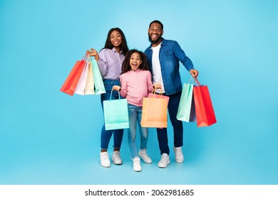 Shopping Fun. Full Body Portrait Of Joyful African American Guy, Lady And Excited Teen Girl Posing With Paper Shopper Bags, Standing Together On Blue Studio Background, Enjoying Sales And Purchases
