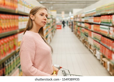 Shopping at the grocery store. A young blonde rolls a cart with products and looks at the shelves of the store. In the background, long rows of shelves with goods. Close up - Shutterstock ID 1549389935