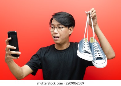 Shopping From Home Concept. A Concept Of Online Shopping Illustrate With Asian Man Holding Phone And Shoes With Happy Expression Isolated Over Red. Man Selling And Buying Stuff In E-commerce