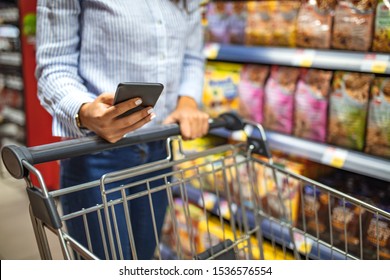 Shopping List. Woman With Smartphone In Store. Grocery Shopping. Gadgets And Shopping.  Picture Of Young Cheerful Woman In Supermarket With Shopping Trolley Choosing Products And Using Phone.