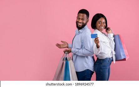 Shopping for lovers day concept. Positive black woman with boyfriend holding gift bags and credit card, posing and looking at camera on pink studio background, banner design with copy space