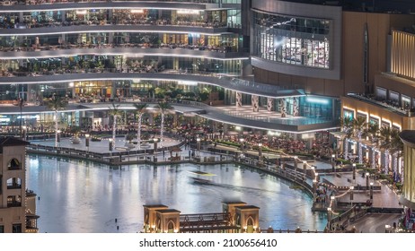 Shopping Mall Exterior With Reataurants Near Fountain In Dubai Downtown Aerial Night Timelapse, United Arab Emirates. Illuminated Bridge And Boats Flouting On A Water