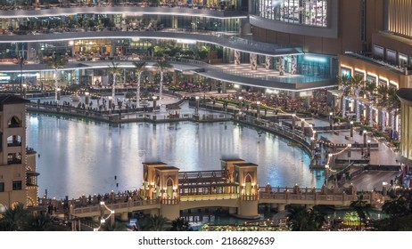 Shopping Mall Exterior With Reataurants Near Fountain In Dubai Downtown Aerial Night Timelapse, United Arab Emirates. Illuminated Bridge And Boats Flouting On A Water