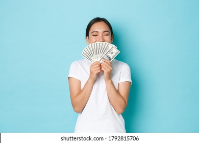 Shopping, Money And Finance Concept. Young Successful And Lucky Cheerful Asian Girl Smelling Dollars And Smiling Satisfied, Enjoying Being Rich, Holding Cash Over Light-blue Background