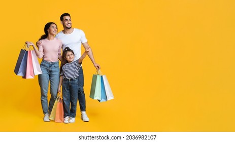 Shopping Offer. Smiling Arab Family With Little Daughter Carrying Shopper Bags And Pointing Aside At Copy Space Over Yellow Background, Happy Parents And Female Kid Enjoying Seasonal Sales, Panorama