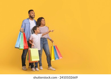 Shopping Offer. Smiling Black Family With Little Son Carrying Shopper Bags And Pointing Aside At Copy Space Over Yellow Background, Happy Parents And Male Kid Enjoying Seasonal Sales, Panorama - Powered by Shutterstock