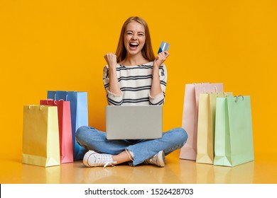 Shopping Online. Joyful Girl Holding Credit Card And Sitting With Laptop On Laps, Surrounded By Bright Paper Bags Over Orange Studio Background