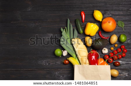 Shopping paper bag with different groceries on dark wooden background, flat lay. Space for text