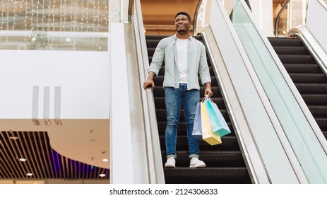 Shopping And Retail Concept. Smiling African American man holding shopper bags, cheerful casual guy going down on escalator in modern mall center and looking away, banner panorama, free copy space