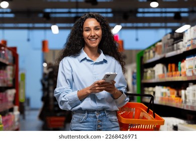 Shopping in a supermarket. A young Latin American woman stands between rows of shelves with a cart. He holds the phone, has a list, writes messages, orders and pays online. Smiling at the camera. - Powered by Shutterstock