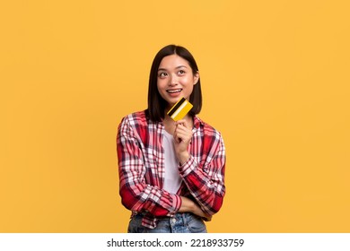 Shopping Time Concept. Dreamy Asian Lady Holding Credit Card And Looking Away, Thinking What To Buy, Posing Over Yellow Studio Background, Copy Space