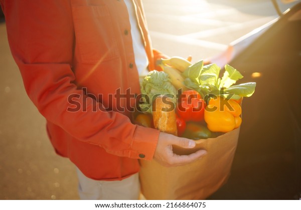 Shopping time. Healthy vegan vegetarian food in a paper\
bag in the male hands. Young man with shopping bag full of\
vegetables near the car.\
