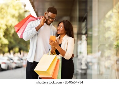 Shopping Together Concept. Portrait Of Happy African American Couple Using Smartphone, Lady Showing Gadget, Guy Holding Shopping Paper Bags, Walking Outdoors. Retail, Sale, Technology