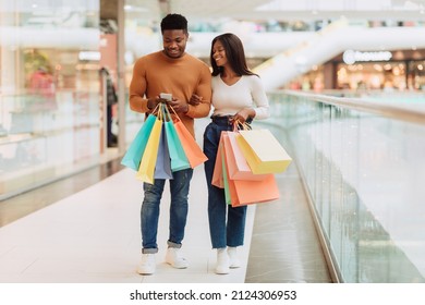 Shopping Together Concept. Portrait of happy African American couple using smartphone, sharing gadget, holding shopper paper bags, walking in city mall center, full body length, free copy space