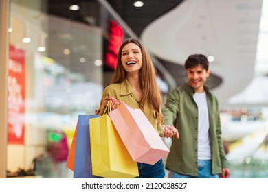 Shopping Together. Joyful Wife Carrying Shopper Bags Pulling Husband Holding His Hand And Laughing Enjoying Sales Weekend In Modern Mall. Shopaholism And Commerce. Selective Focus