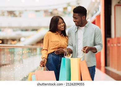 Shopping Together. Portrait of excited happy African American couple spending time together in modern city mall bying gift, holding open colorful bags with purchases and smiling, looking at each other
