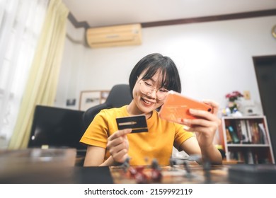 Shopping Via Online Technology Concept. Young Adult Asian Woman Using Smartphone And Credit Card Buy For Hobby. People Lifestyle At Home. Boardgame On Foreground.