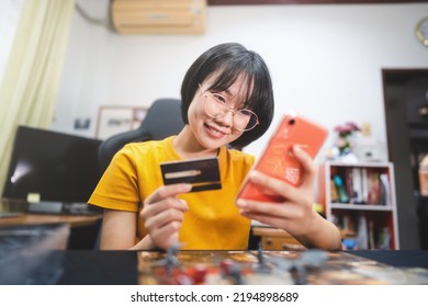 Shopping Via Online Technology Concept. Young Adult Asian Woman Using Smartphone And Credit Card Buy For Hobby. People Lifestyle At Home. Boardgame On Foreground.