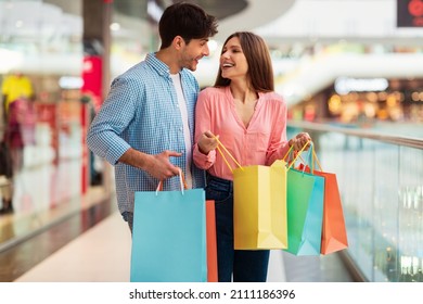 Shopping Weekend. Cheerful Spouses Buying New Clothes During Great Sales Holding Colorful Shopper Bags Smiling To Each Other Standing In Modern Hypermarket. Consumerism Concept