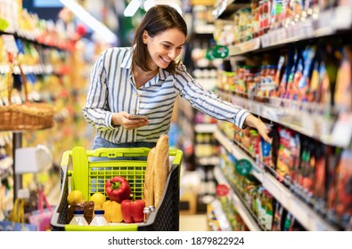 Shopping. Young Smiling Woman Holding And Using Mobile Phone Buying Food Groceries Standing In Supermarket. Female Customer With Smartphone Taking Healthy Products From Shelf At The Shop - Shutterstock ID 1879822924