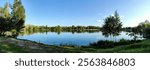 The shore of the lake with deciduous trees, reflections on the water and the blue sky with small clouds on summer evening. The panorama. Russia. Nizhny Novgorod. Lake Svetloyarskoe. August 2024.Photo.