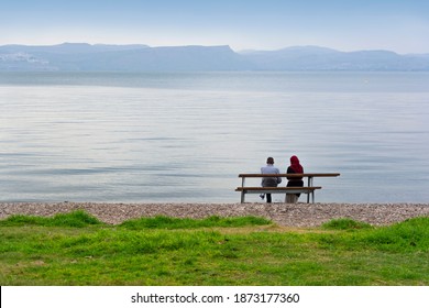 Shore of Lake Kinneret, Israel. A man and a woman in a Muslim headscarf sitting at the water's edge and looking at the lake. - Powered by Shutterstock