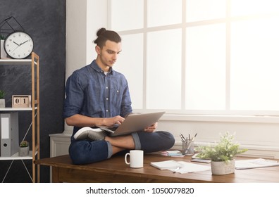 Short break for yoga in office. Flexible man practicing yoga at workplace, sitting on desk in lotus pose and having coffee with laptop, copy space. Active employee at work, healthy lifestyle concept - Shutterstock ID 1050079820