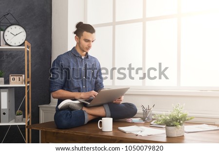 Short break for yoga in office. Flexible man practicing yoga at workplace, sitting on desk in lotus pose and having coffee with laptop, copy space. Active employee at work, healthy lifestyle concept