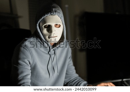 Short of computer hacker wearing white mask stealing data from a laptop. Masked hacker using laptop computer for organizing massive data breach attack on corporate servers in neon room.