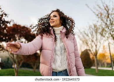 In A Short Down Jacket In The Park Stands A Girl Reaching Out Her Hand Towards Someone. High Quality Photo