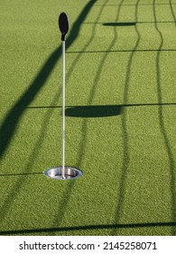 Short flagstick above metallic cup in artificial putting green, with slightly curved afternoon shadows of railing and handrail, on top deck of cruise ship. For themes of golf, practice, recreation.