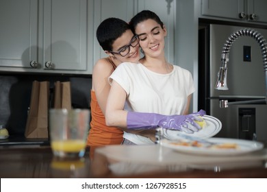 A Short Hair Gay Woman Embracing Her Couple While She Cleans The Dishes. LGBT Women In Everyday Domestic Activities. Home Tasks Cooperative House Cleaning And Relationships