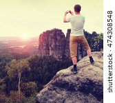 Short hair man on cliff of rock takes photo by smart phone of rocks and landscape bellow. Sunny evening in rocky mountains. Hiker with grey shirt and colorful trekking pants. 