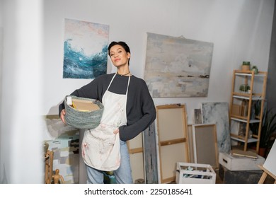 Short haired artist in apron holding basket and equipment near drawings in studio
