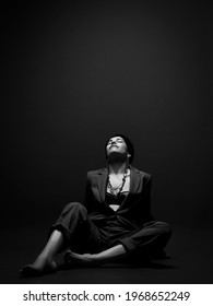Short haired brunette woman in business smart casual suit on naked body sits on floor with head thrown back, feeling pleasure, relax over dark background. Stylish female wear and fashion concept