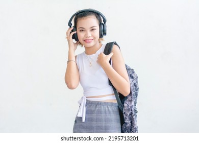 A short haired student wearing a white sleeveless top and printed skirt holding her backpack while listening to music on her wireless headphones. Isolated on a white background. - Shutterstock ID 2195713201