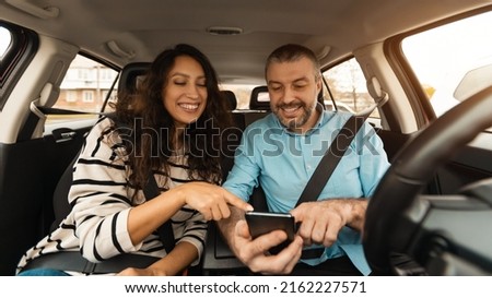 Short Road Route. Portrait of smiling couple sitting inside luxury car pointing fingers at smart phone, cheerful male driver holding cell, showing choosing location, using digital map application