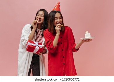 Short-haired Asian Girls In Oversized Shirts Pose In Good Mood And Blow Out Party Horns. Women Hold Piece Of Birthday Cake And Gift Box.