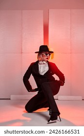 short-haired dancer posing while sitting in studio with red backlit background. young caucasian female in black suirt, sunglasses, hat alone. portrait. grace, confidence, people, dance