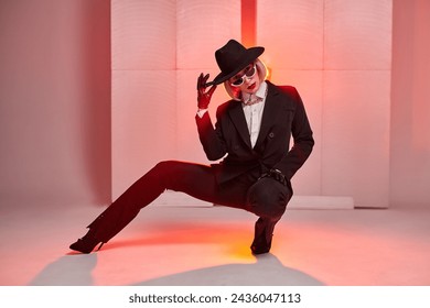 short-haired dancer posing while sitting in studio with red backlit background. young caucasian female in black suirt, sunglasses, hat alone. portrait. grace, confidence, people, dance